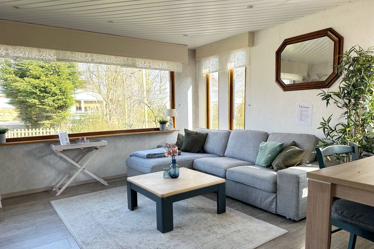A modern living room with a grey sectional sofa, a wooden coffee table, and a large window offering a view of trees and a white fence.