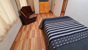 A small, compact room with a wooden floor, a bed with striped bedding, a brown armchair, a wooden dresser, and a television on a stand. - Ukumary Gosth (Cusco)