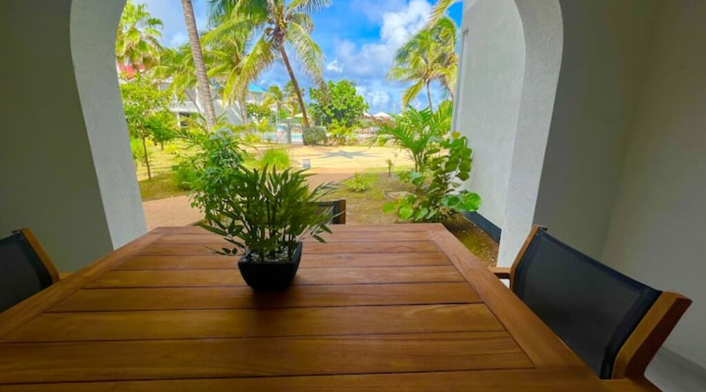 A wooden table with chairs, a potted plant, and a view of a tropical garden with palm trees and a clear sky.
