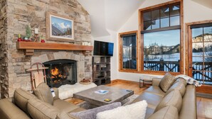 A cozy living room with a stone fireplace, a large window with a view of a snowy landscape, and a comfortable beige sofa. - Ski-in/out luxury condo. Best in the complex. Outdoor pool and hot tubs!! (Bachelor Gulch)