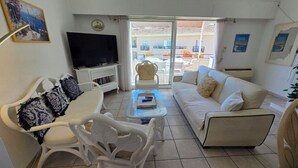 A living room with a sofa, armchair, TV, and a glass door leading outside. - Appartement 3 Pièce Climatisé, Centre Ville de Sainte-maxime (Sainte-Maxime)