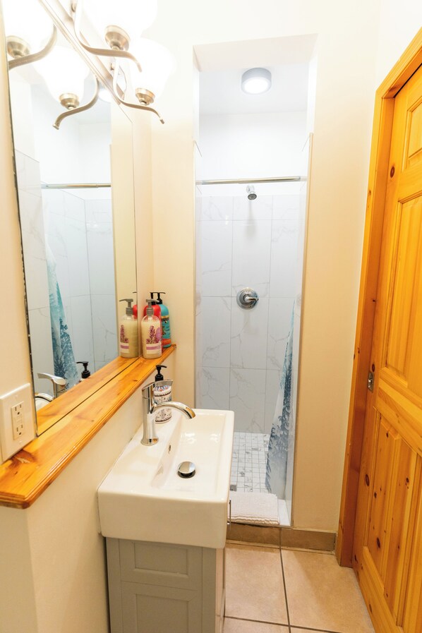 A bathroom with a white sink, mirror, and shower. There are toiletries on the sink and a wooden door to the right.