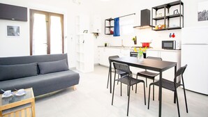 Moderni asunto, jossa on harmaa sohva, ruokapöytä neljällä tuolilla, valkoisilla kaapeilla varustettu keittiö, mikroaaltouuni ja punainen kattila liedellä.