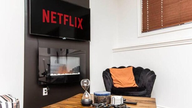 Una habitación con un televisor montado en la pared que muestra 'NETFLIX', un sillón de cuero negro con una manta naranja, una mesa de madera con un reloj, una taza y un control remoto.