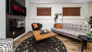 Una sala de estar con un sofá gris, una mesa de centro de madera, un sillón negro y un televisor montado en la pared mostrando 'NETFLIX'.