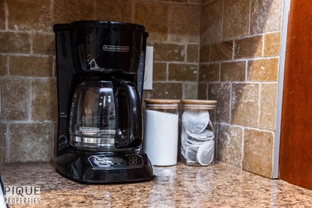 A black coffee maker on a granite countertop with a stone backsplash.
