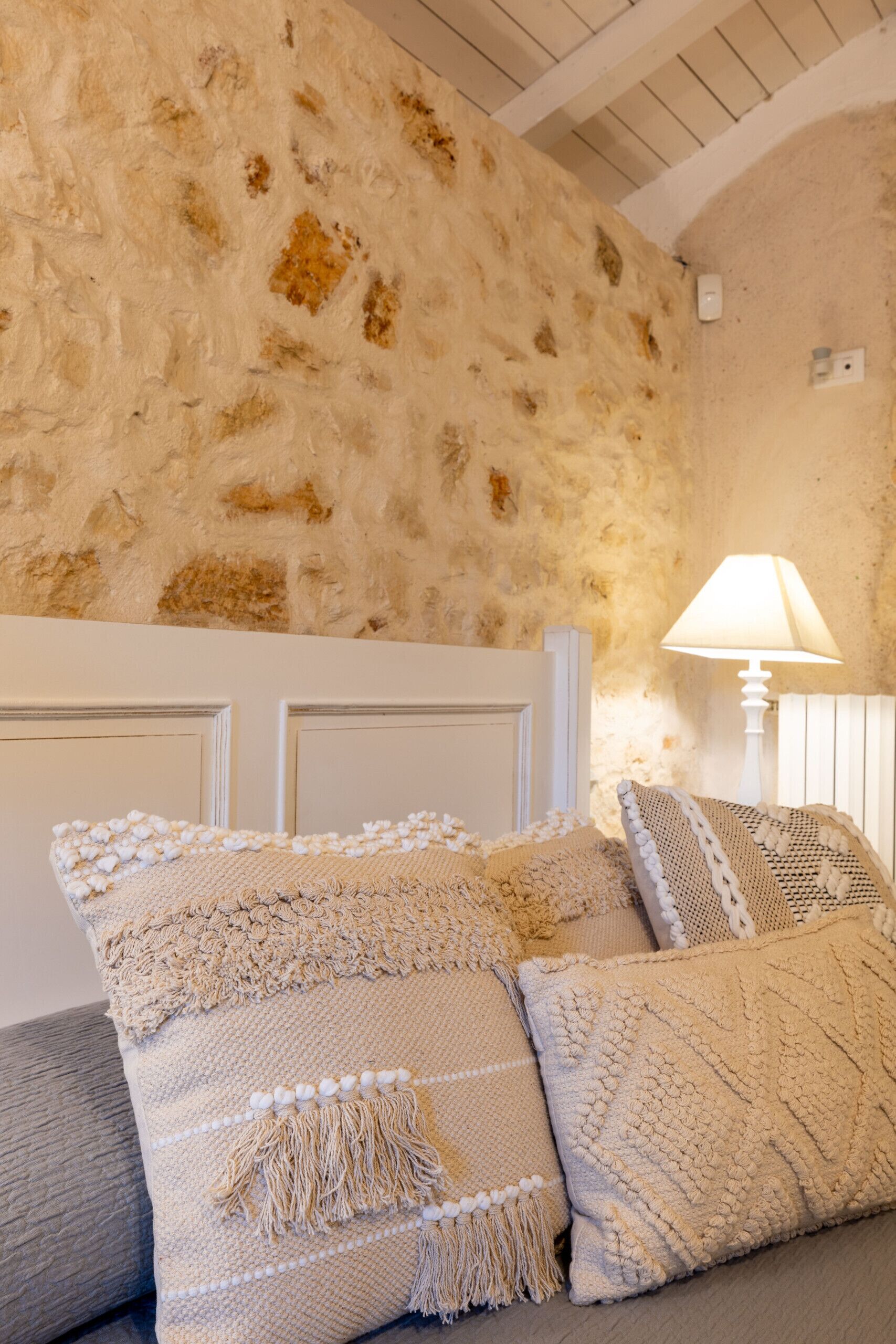 Une pièce avec un mur en pierre, une tête de lit blanche et une lampe sur une table de chevet.