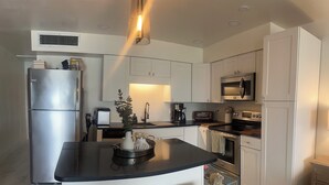 Une cuisine moderne avec des armoires blanches, un réfrigérateur en acier inoxydable, un four à micro-ondes et une cuisinière. On y trouve deux tabourets avec des assises en osier et un petit îlot central agrémenté d’une plante.