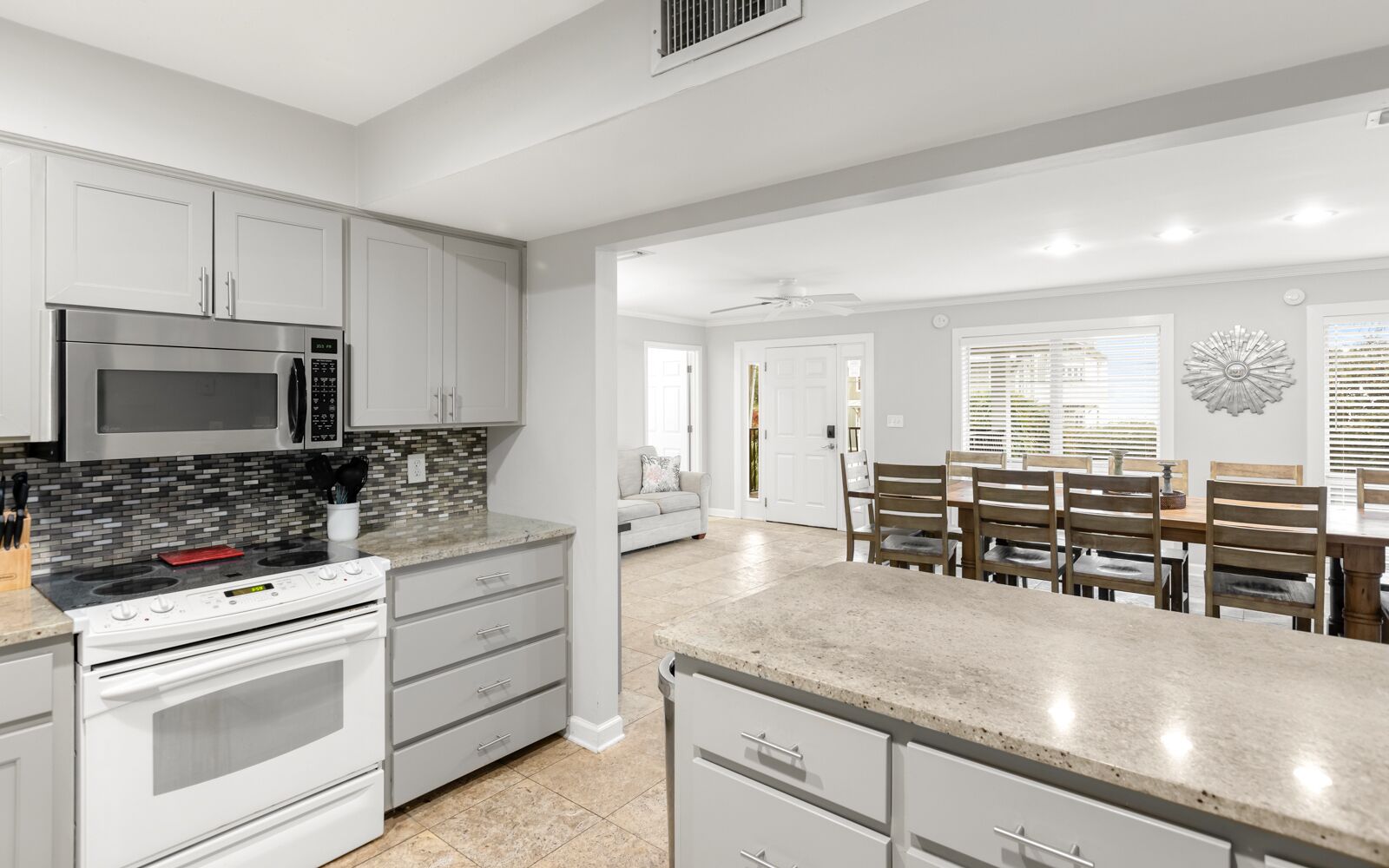 A modern kitchen with white cabinetry, a stainless steel microwave, and a built-in oven. There is a dining area with wooden chairs and a table.