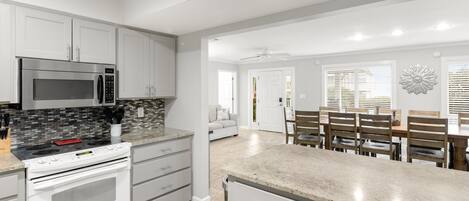 A modern kitchen with white cabinetry, a stainless steel microwave, and a built-in oven. There is a dining area with wooden chairs and a table.