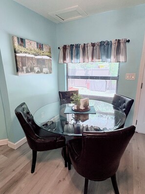 A dining area with a glass table surrounded by leather chairs, a window with blinds, and a wall-mounted picture. - Vibrant Bungalow in Historical Fort Myers District (Fort Myers)