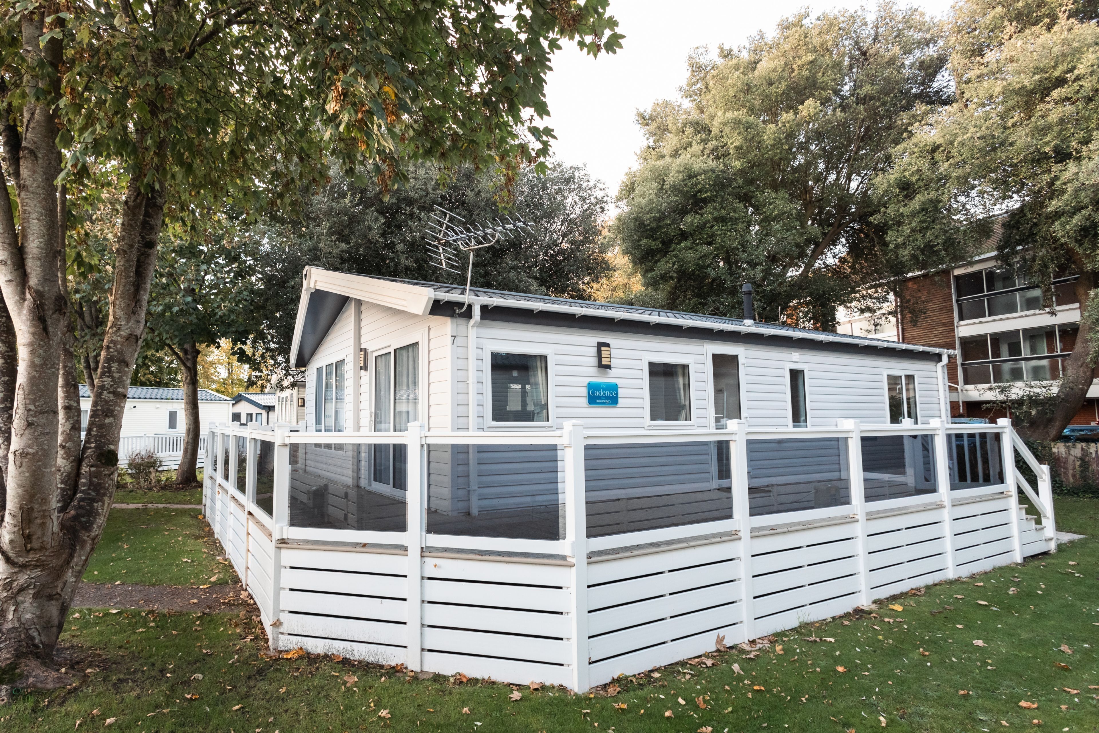 A white mobile home with a fenced deck, surrounded by trees and a grassy area.
