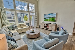A living room with a sofa, armchairs, a coffee table, a TV, and a view of a house through large windows. - Bayside * 4BR Luxury House (Selbyville)
