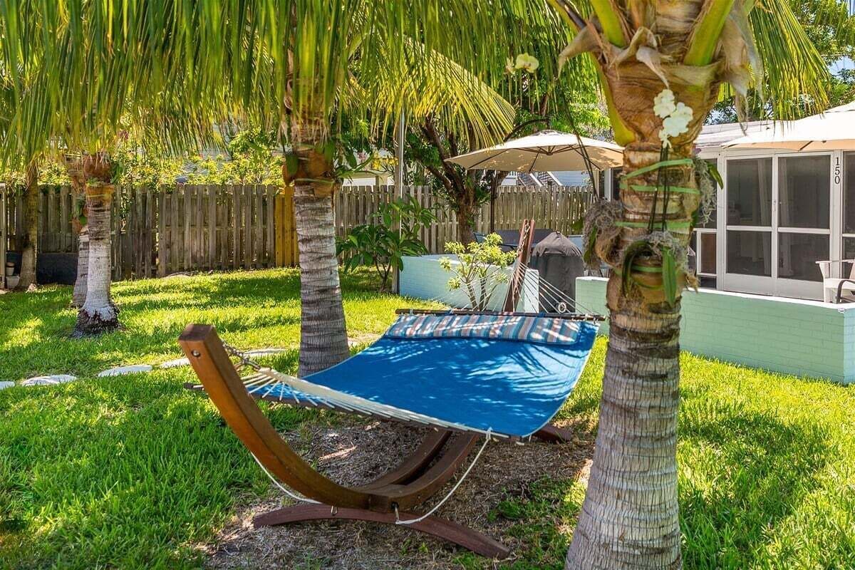 Eine Hängematte unter einer Palme im Garten mit Holzzaun, Grill und überdachter Terrasse.