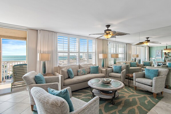 A spacious living room with a large window overlooking the beach, a ceiling fan, and a dining area with a view.
