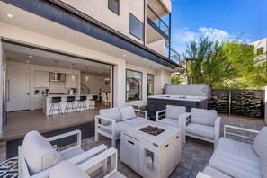 A modern outdoor living area with a hot tub, white furniture, and a kitchen visible through an open door. - Desert Chic at Desert Color by Ember Stays (St. George)