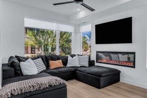 A modern living room with a sectional sofa, a fireplace, and large windows offering a view of greenery. - Desert Chic at Desert Color by Ember Stays (St. George)