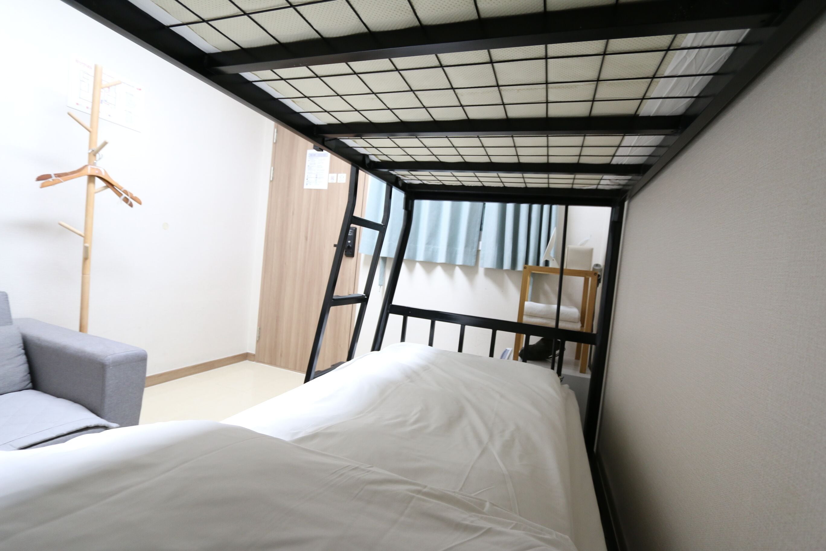 Une petite pièce avec un lit superposé, un canapé gris et un porte-manteau en bois.