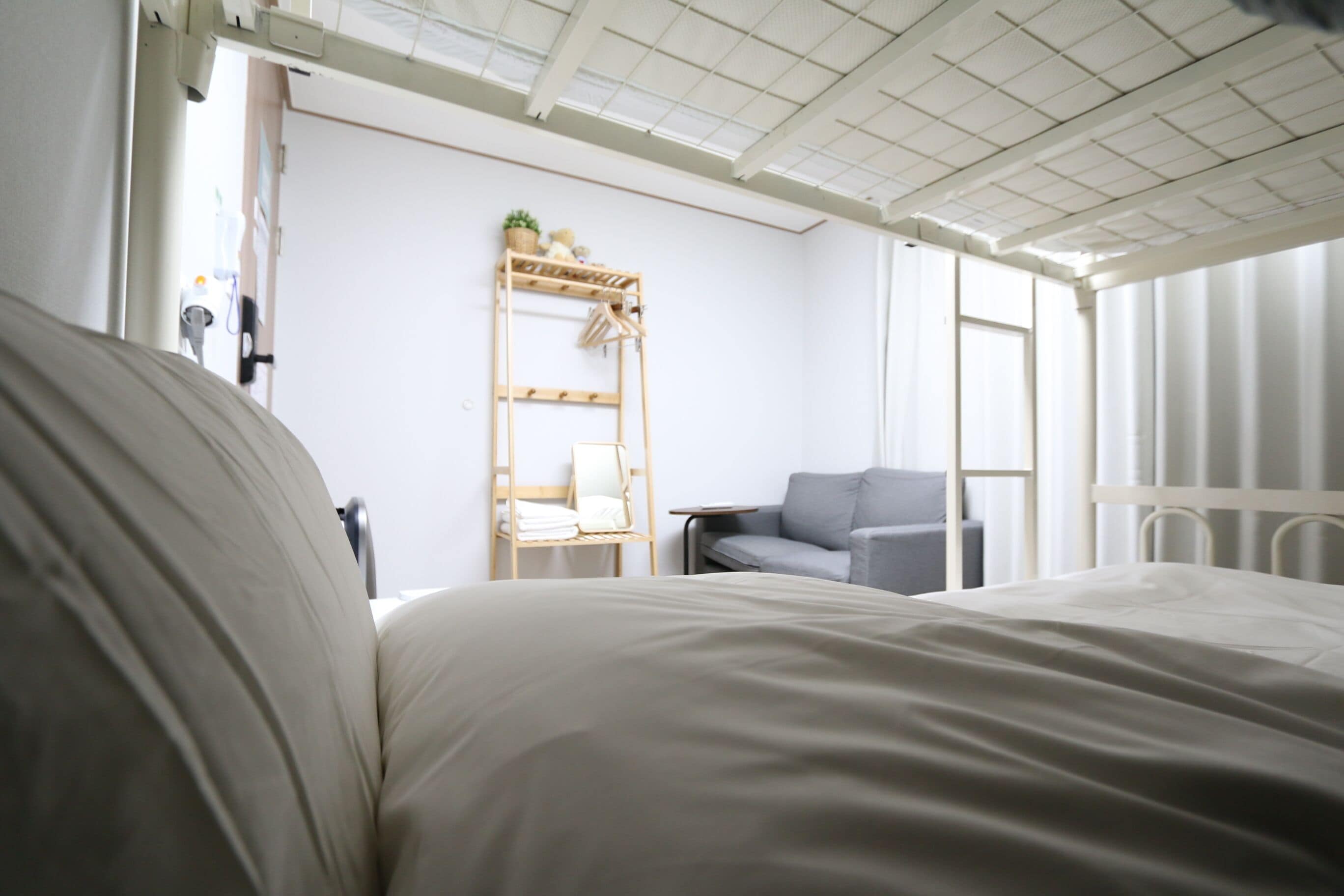 A bedroom with a bed, a grey armchair, a wooden shelf with towels, and a window.