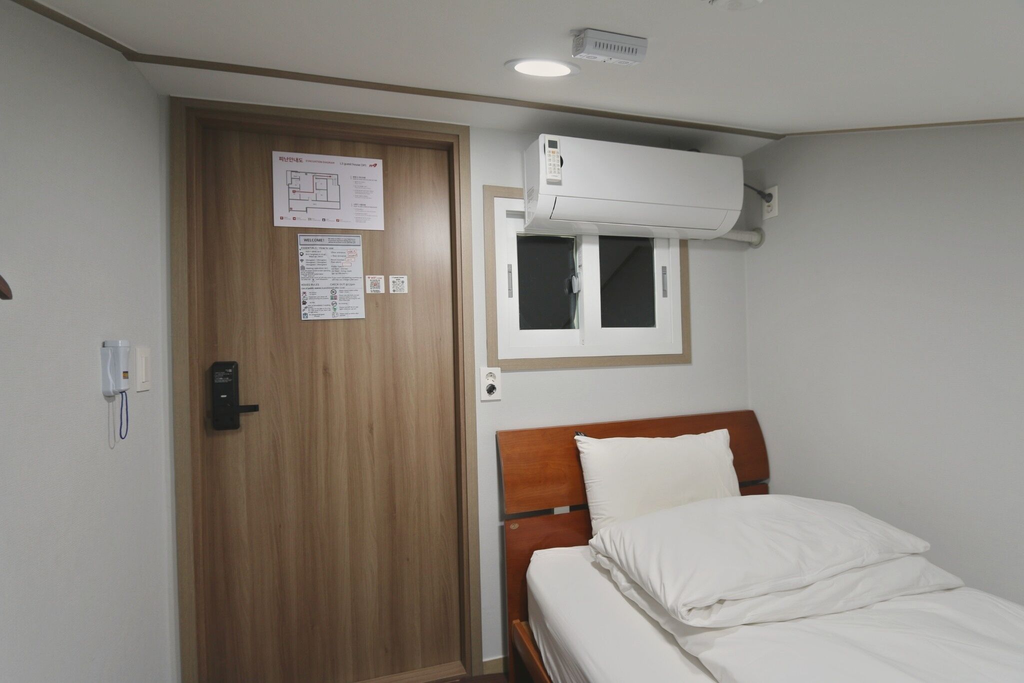 A hotel room with a wooden door, a bed with white bedding, an air conditioning unit mounted on the wall, and a small window.
