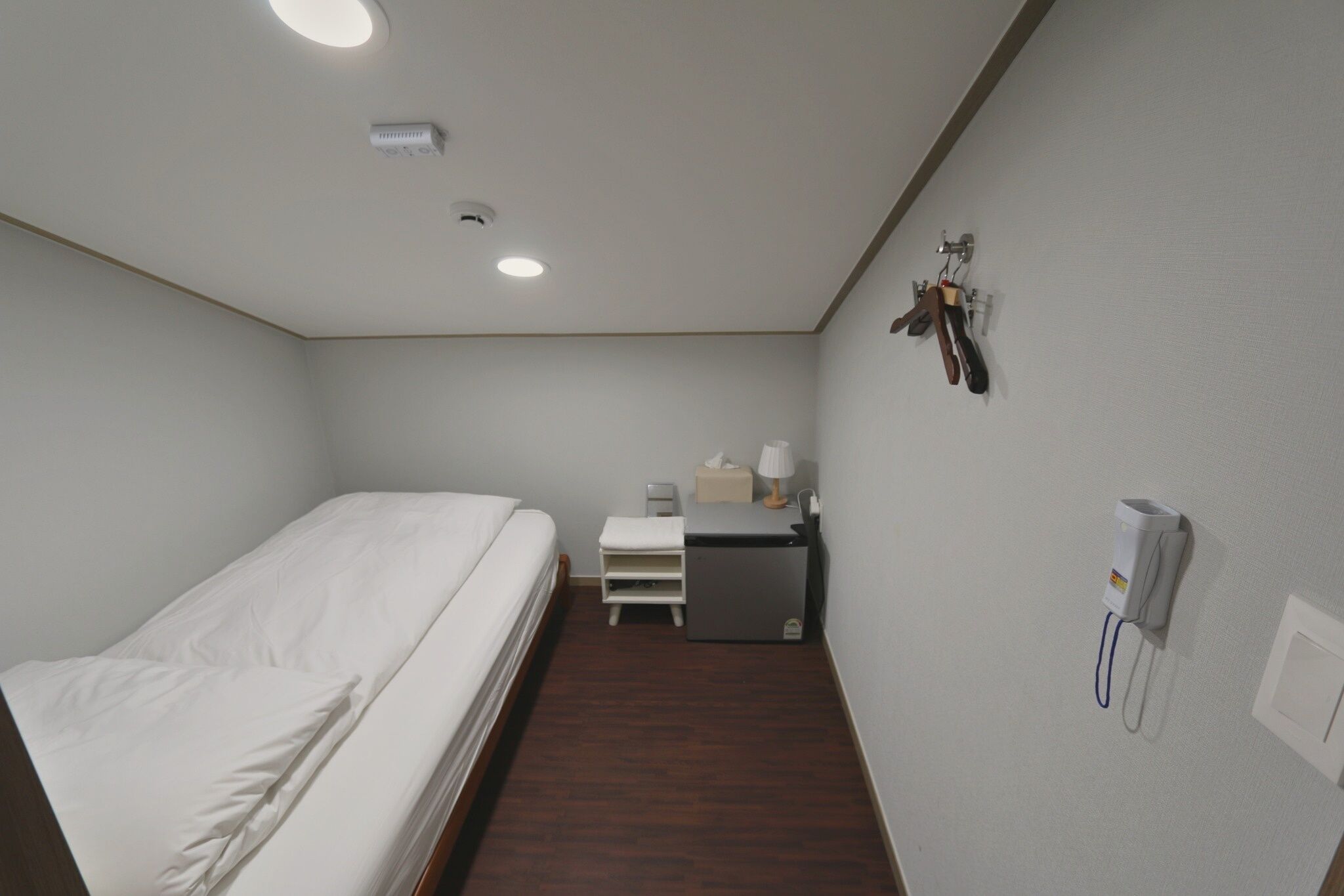 A small hotel room with a single bed, a desk with a lamp, a small table, and a wall-mounted phone.