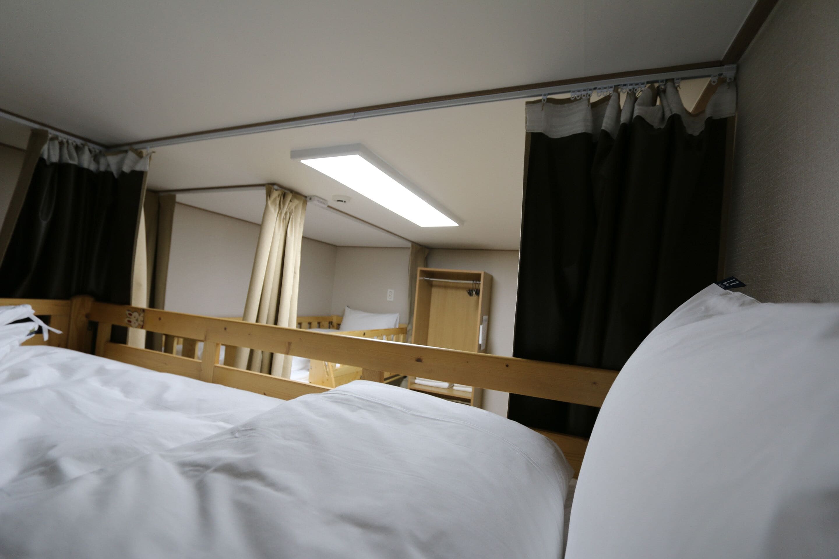 A single bed with a wooden headboard, a bedside table, and a mirror reflecting a room with a window, curtains, and a ceiling light.