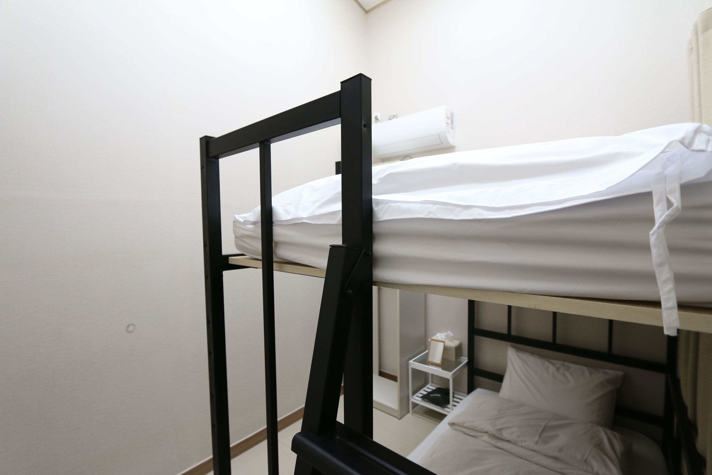 Ein Stockbett mit weißer Bettwäsche, ein kleiner Nachttisch mit Taschentuchbox und eine weiße Wand.