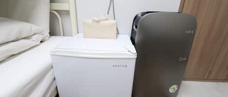 A compact, white portable air cooler with a beige tissue box on top, placed on a wooden floor next to a bed with white bedding.