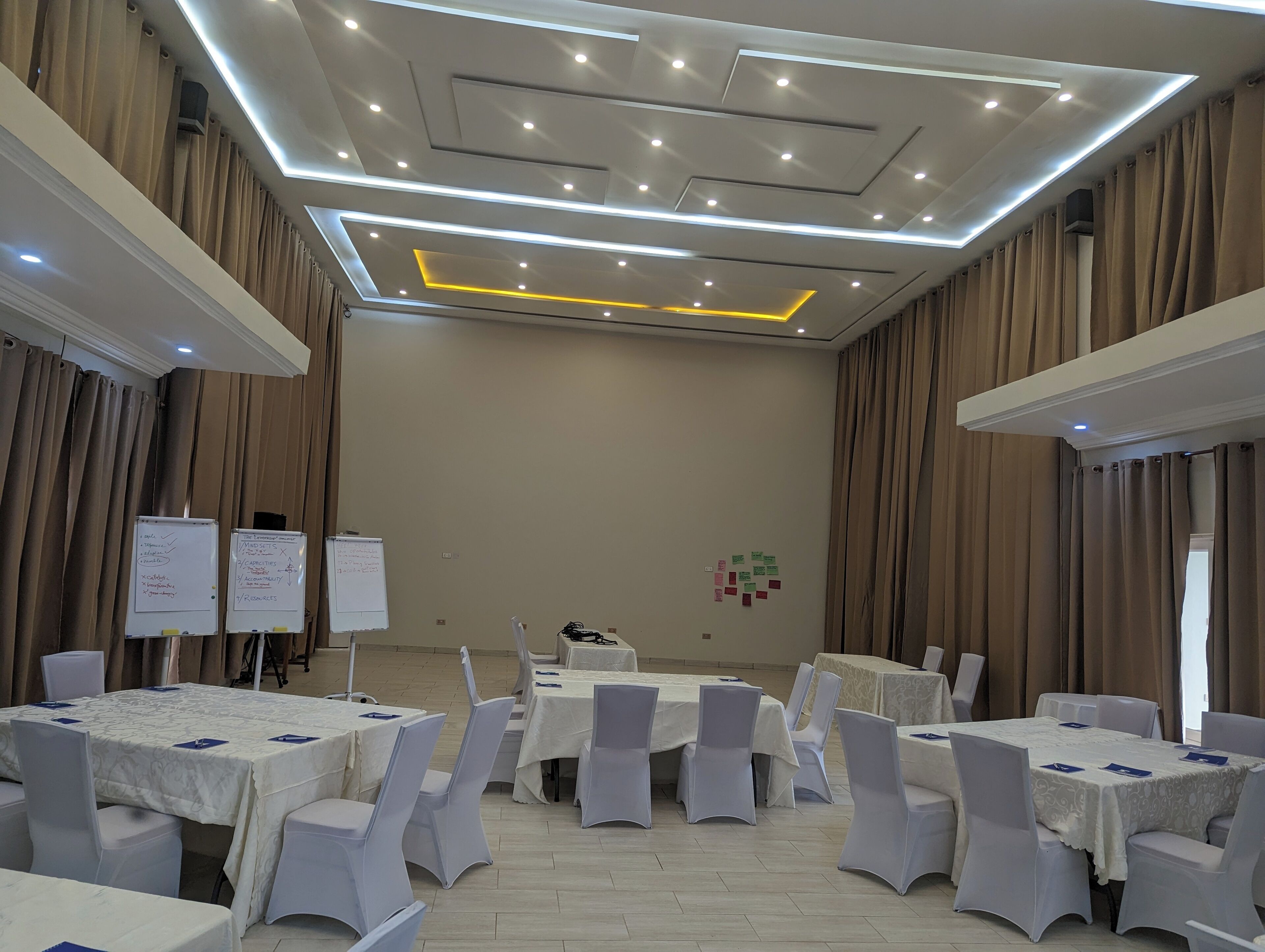 Un salón de conferencias con mesas y sillas blancas, una pizarra y un diseño de techo moderno.