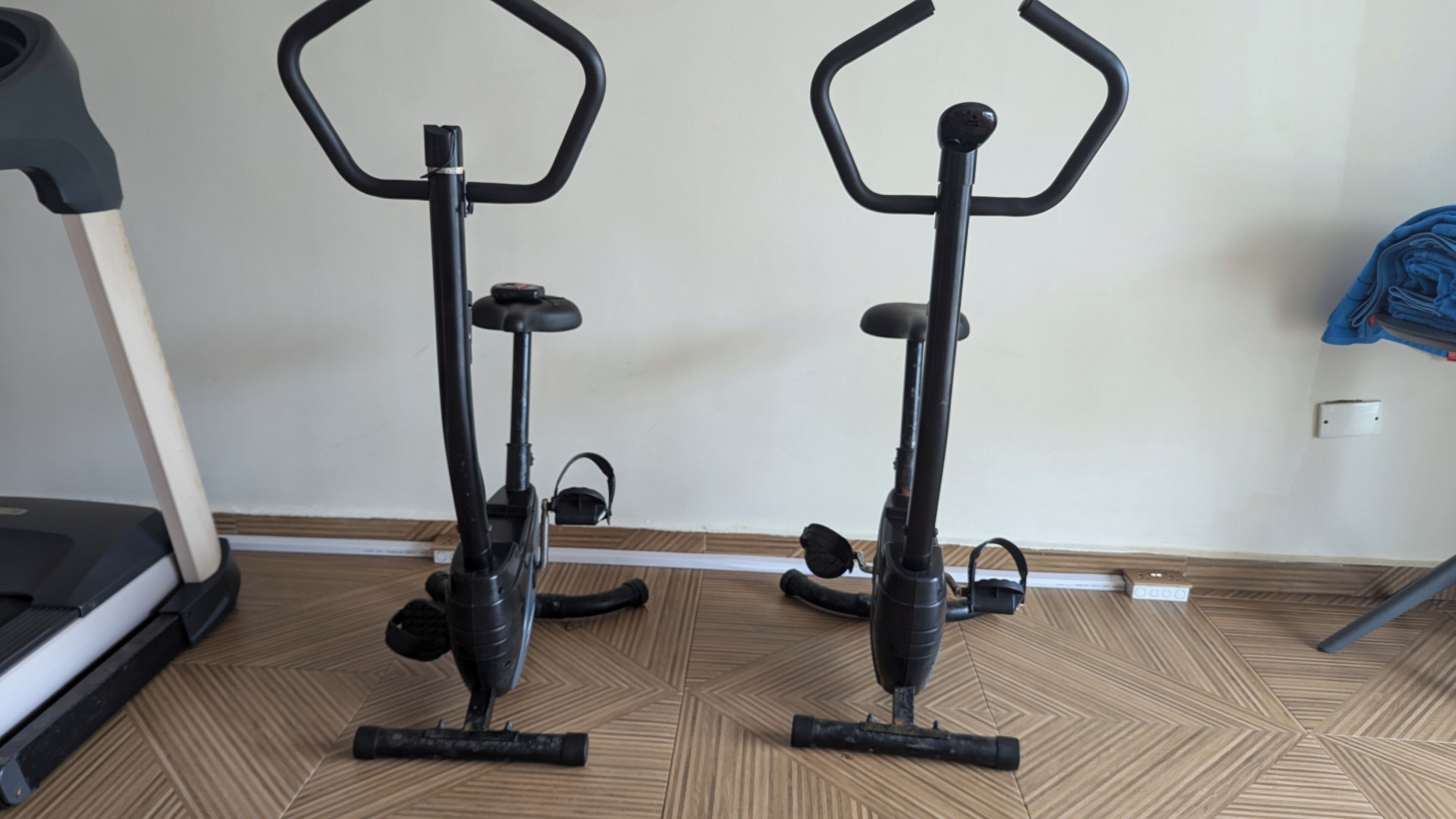Dos bicicletas estáticas en una sala con piso estampado, una caminadora y una toalla azul sobre una silla.