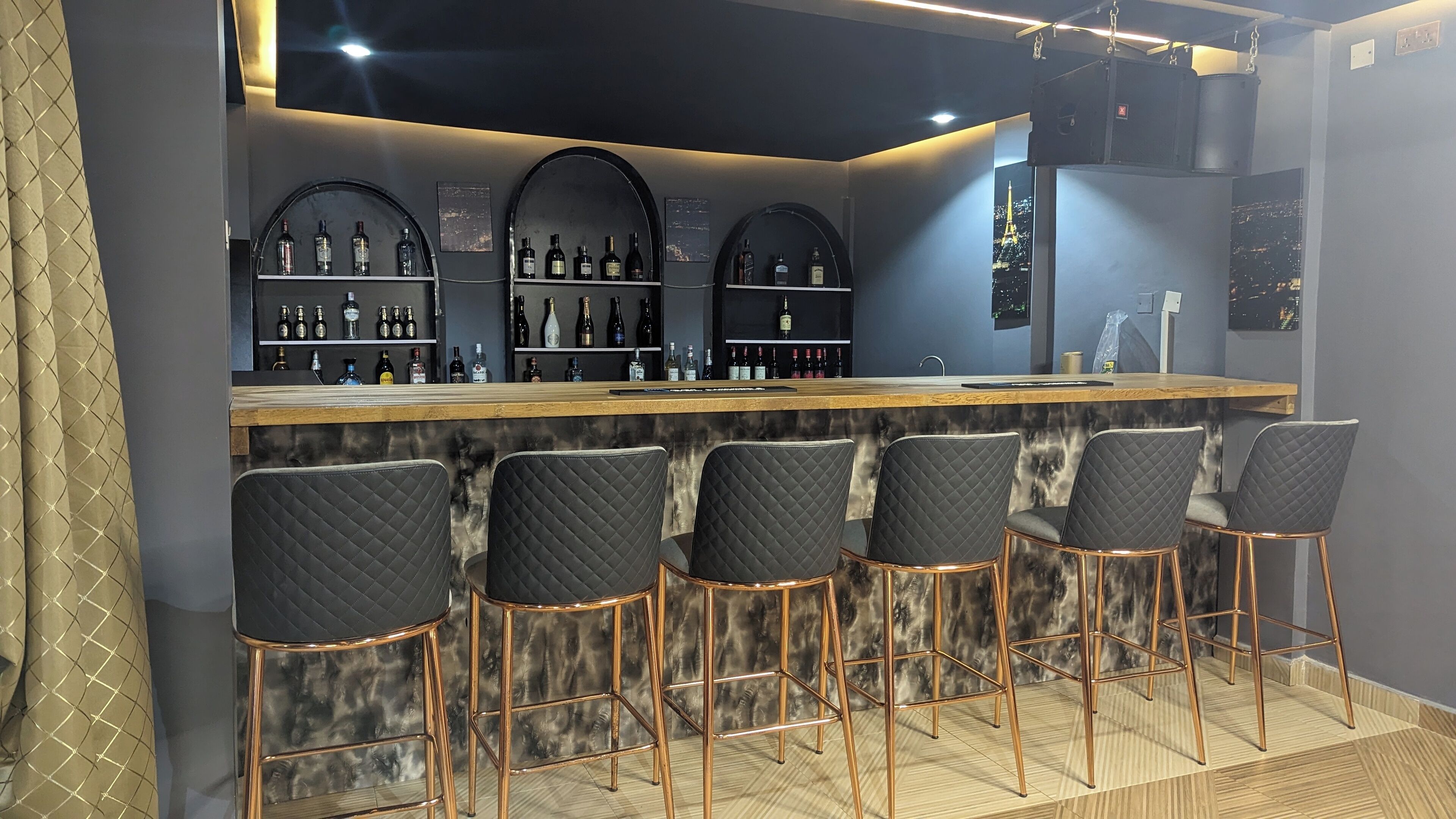 Un espace bar doté d’un comptoir en marbre, de tabourets de bar noirs rembourrés et d’étagères remplies de bouteilles d’alcool.