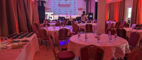 A conference room with round tables, chairs, and a stage with a large screen displaying the event name.