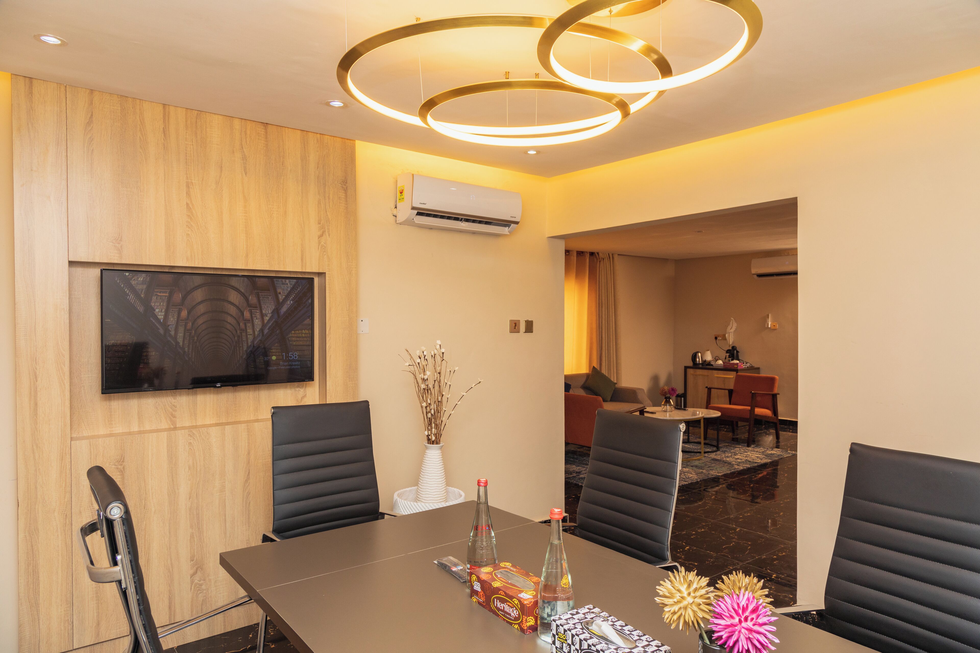 A modern hotel room with a wooden panel wall, a flat-screen TV, a dining table with chairs, and a decorative vase.