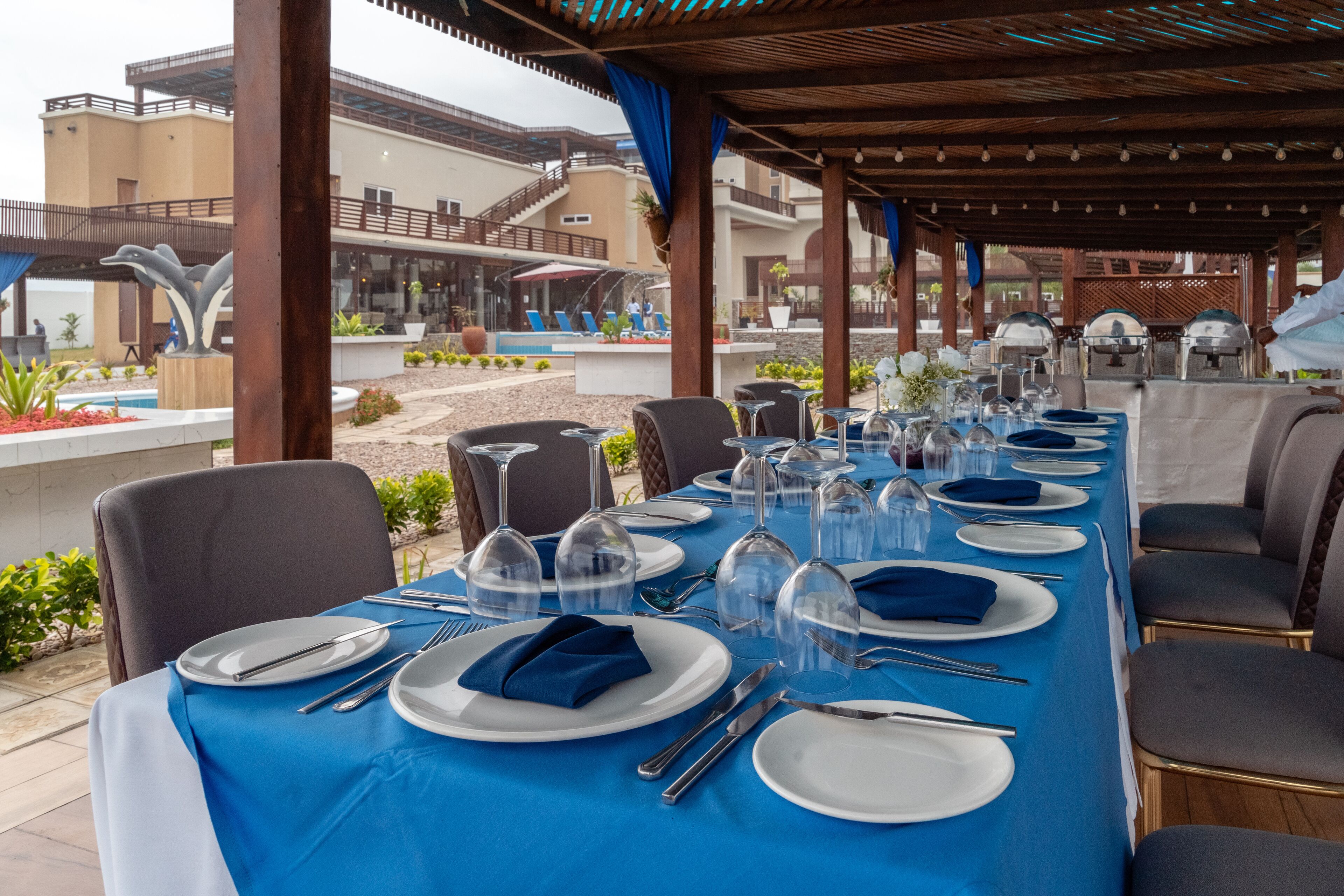 Una larga mesa de comedor dispuesta con platos blancos, servilletas azules y cristalería transparente, bajo una pérgola de madera con vista a una piscina y edificios.