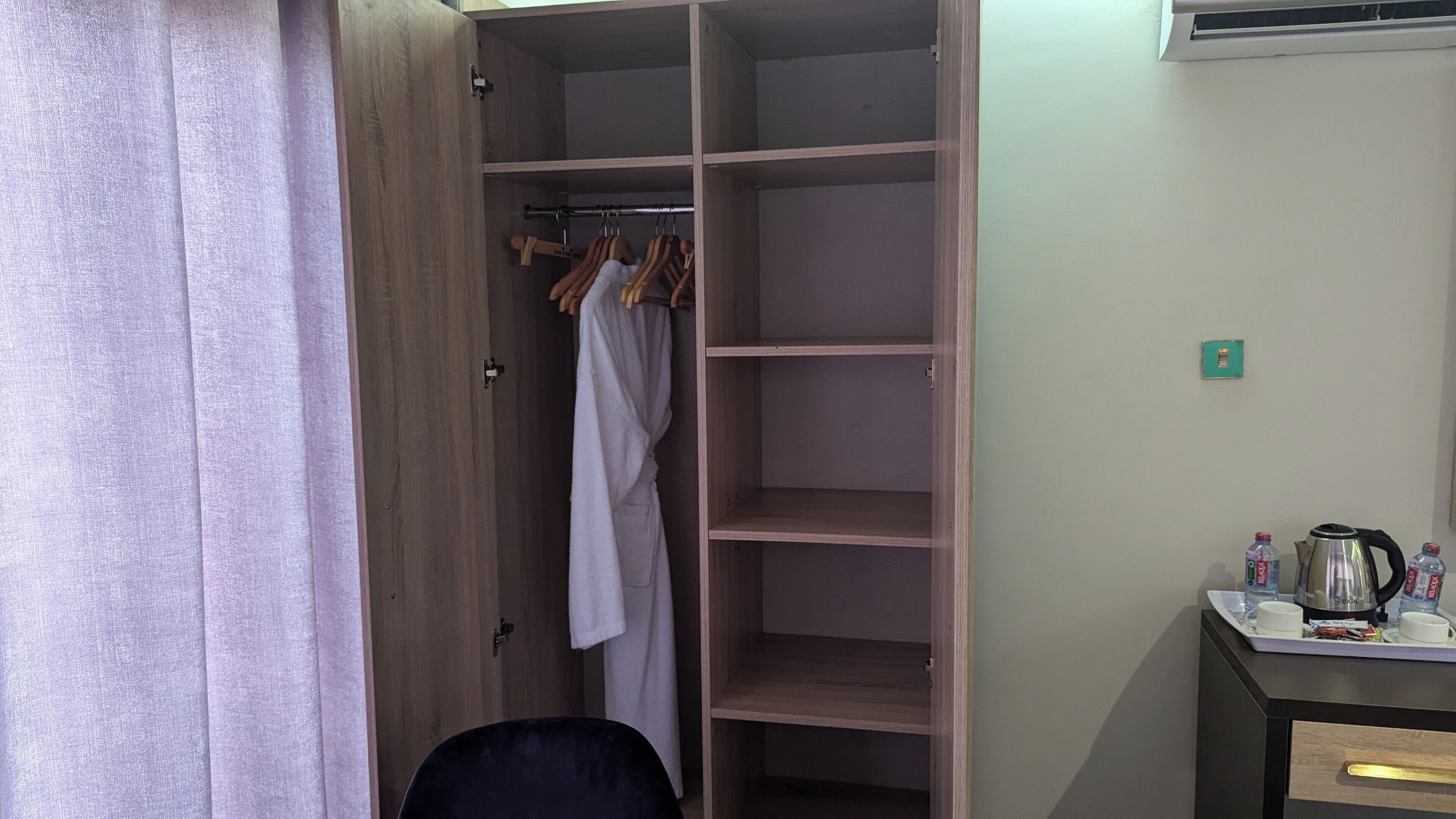 A room with a wooden wardrobe, a small desk with a kettle and cups, and a chair.