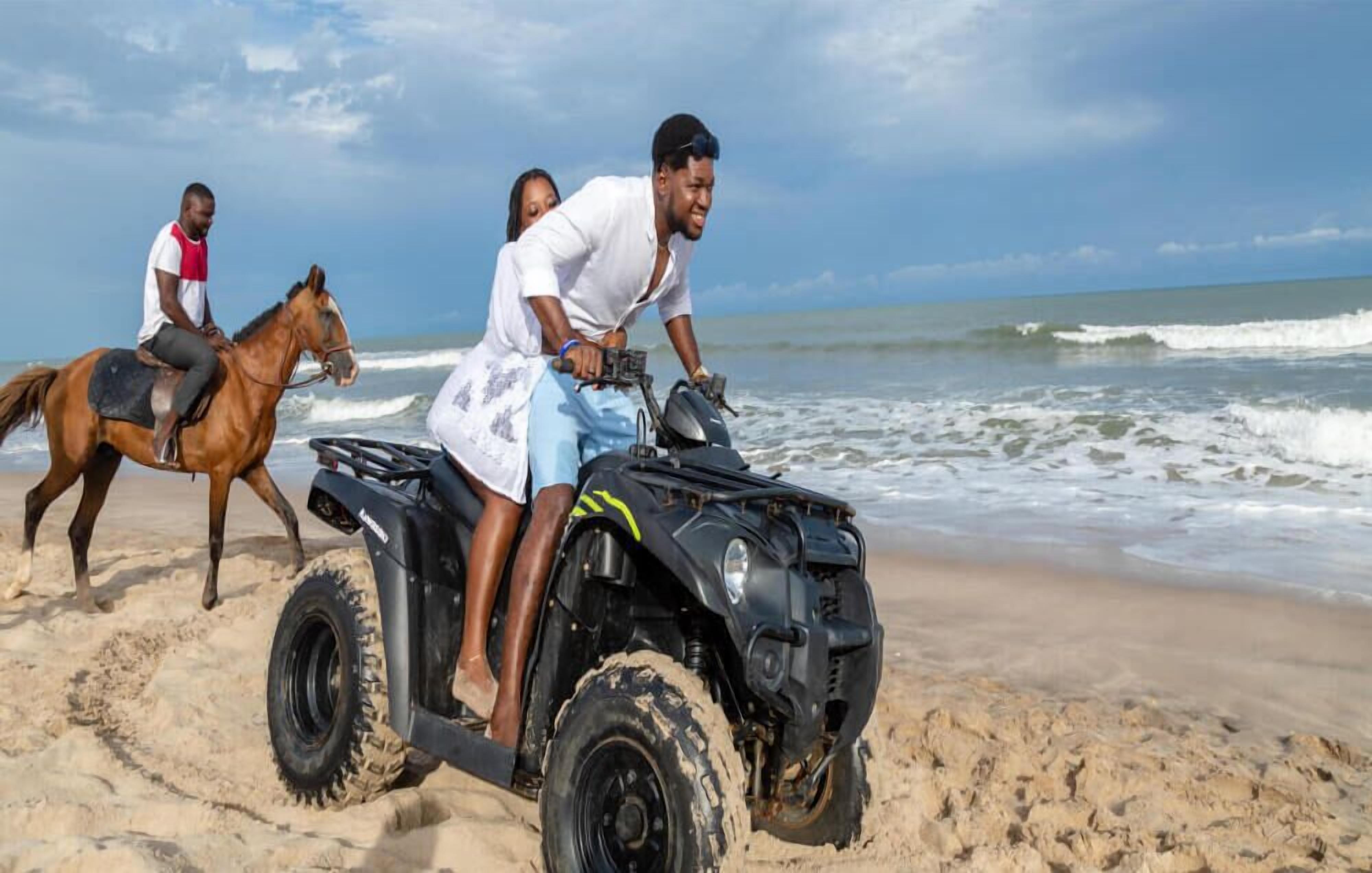 A beach scene with a person riding a horse and another on a four-wheeled vehicle.