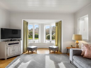 A living room with a television, two ottomans, a sofa, a lamp, and a view through large windows.