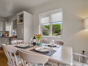 A dining area with a table set for four, white chairs, a vase with flowers, and a bottle of wine. - An tSean Scoil, Clifden (Clifden)
