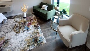 Un salon moderne avec un canapé vert, un fauteuil blanc et une table basse. Deux tableaux encadrés ornent le mur, une grande fenêtre offre une vue dégagée et un radiateur se trouve sous le canapé.