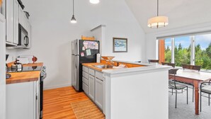 A modern kitchen with white cabinetry, a wooden countertop, and a built-in island with a sink. There are pendant lights, a refrigerator with magnets, and a dining area with a table and chairs. - Saddleback - Rock Pond Condo 43 - ski in/out, pet friendly! (Rangeley)