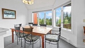A dining area with a wooden table and metal chairs, large windows with a view of trees, and a ceiling light fixture. - Saddleback - Rock Pond Condo 43 - ski in/out, pet friendly! (Rangeley)