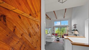 A modern interior with a wooden ceiling, a white sofa set, and a large window offering a view of greenery. - Saddleback - Rock Pond Condo 43 - ski in/out, pet friendly! (Rangeley)