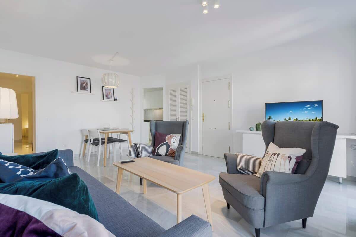 Een moderne woonkamer met een bank, fauteuil en salontafel. Er is een eethoek met een tafel en stoelen, en een keuken met kasten en een koelkast.