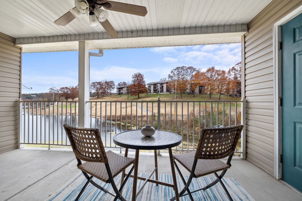 A covered porch with two wicker chairs and a small table, overlooking a residential area with trees and a building in the background.