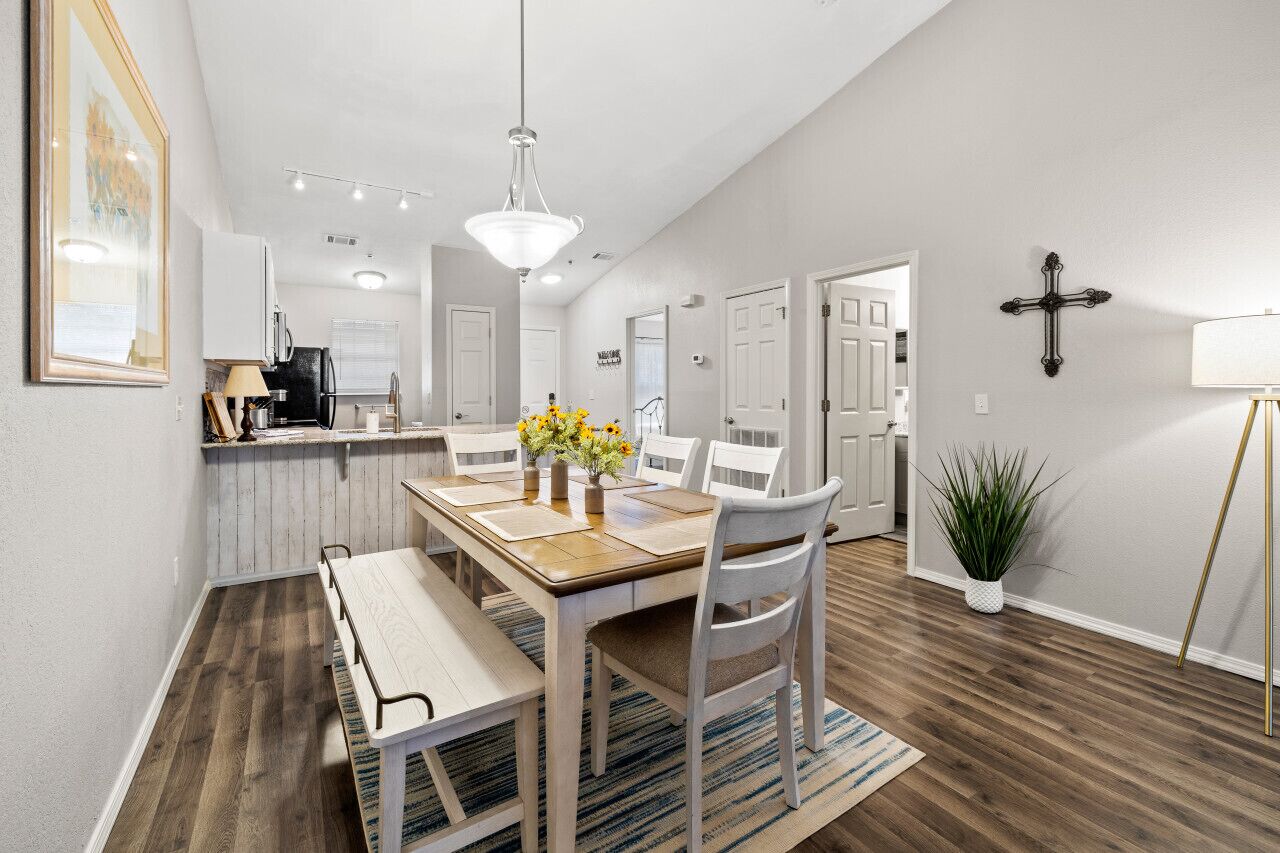 A dining area with a wooden table, white chairs, and a chandelier. There is a kitchen in the background with white cabinets and a refrigerator.