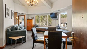 A cozy living room with a sofa, a dining table with chairs, a television, and a large window with a view of trees and houses. - 321 | Luxe Top Floor 1BD - A/C, Pool & Kitchen (Olympic Valley)