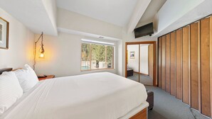 A bedroom with a large bed, a bedside lamp, a window with a view of trees, and a wooden wardrobe. - 321 | Luxe Top Floor 1BD - A/C, Pool & Kitchen (Olympic Valley)