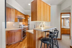 Une cuisine moderne avec des meubles en bois, un réfrigérateur en acier inoxydable, un four encastré et un coin bar avec des tabourets noirs.