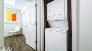 Una lavandería con una secadora y una lavadora blancas, una silla blanca y una puerta que da a otra habitación.