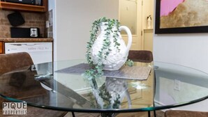 Una mesa de vidrio con una jarra blanca y plantas verdes, una silla de cuero marrón y una cocina con microondas y tostadora.